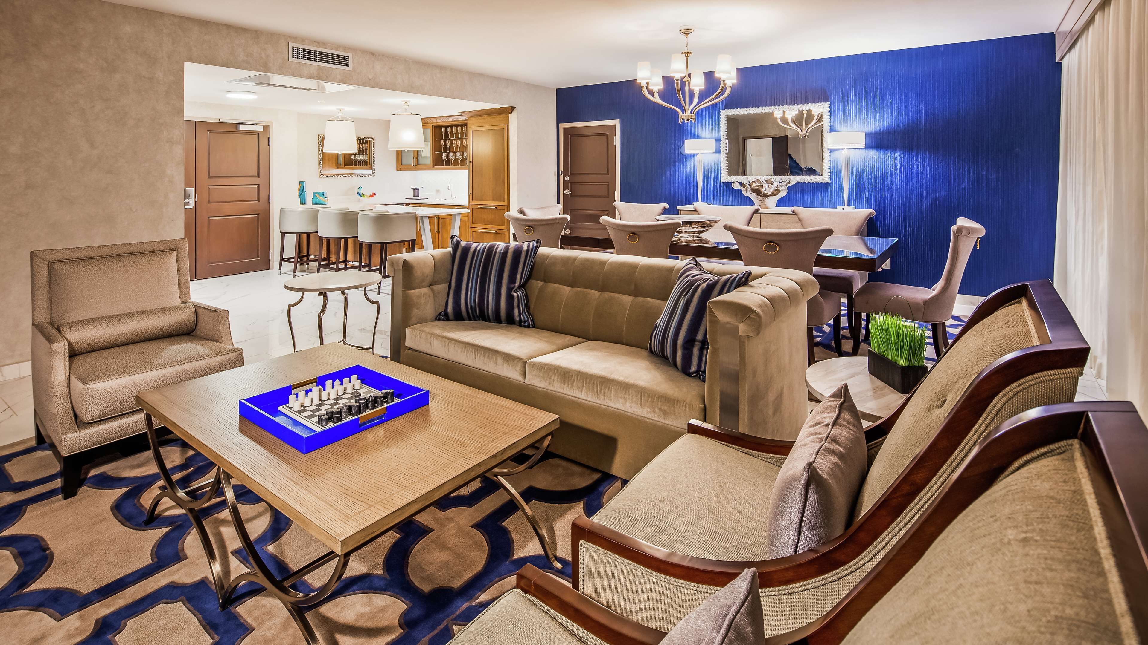 A hotel room with a living area featuring a sofa, armchairs, a coffee table with a chessboard, a dining area with a table and chairs, and a kitchen in the background.