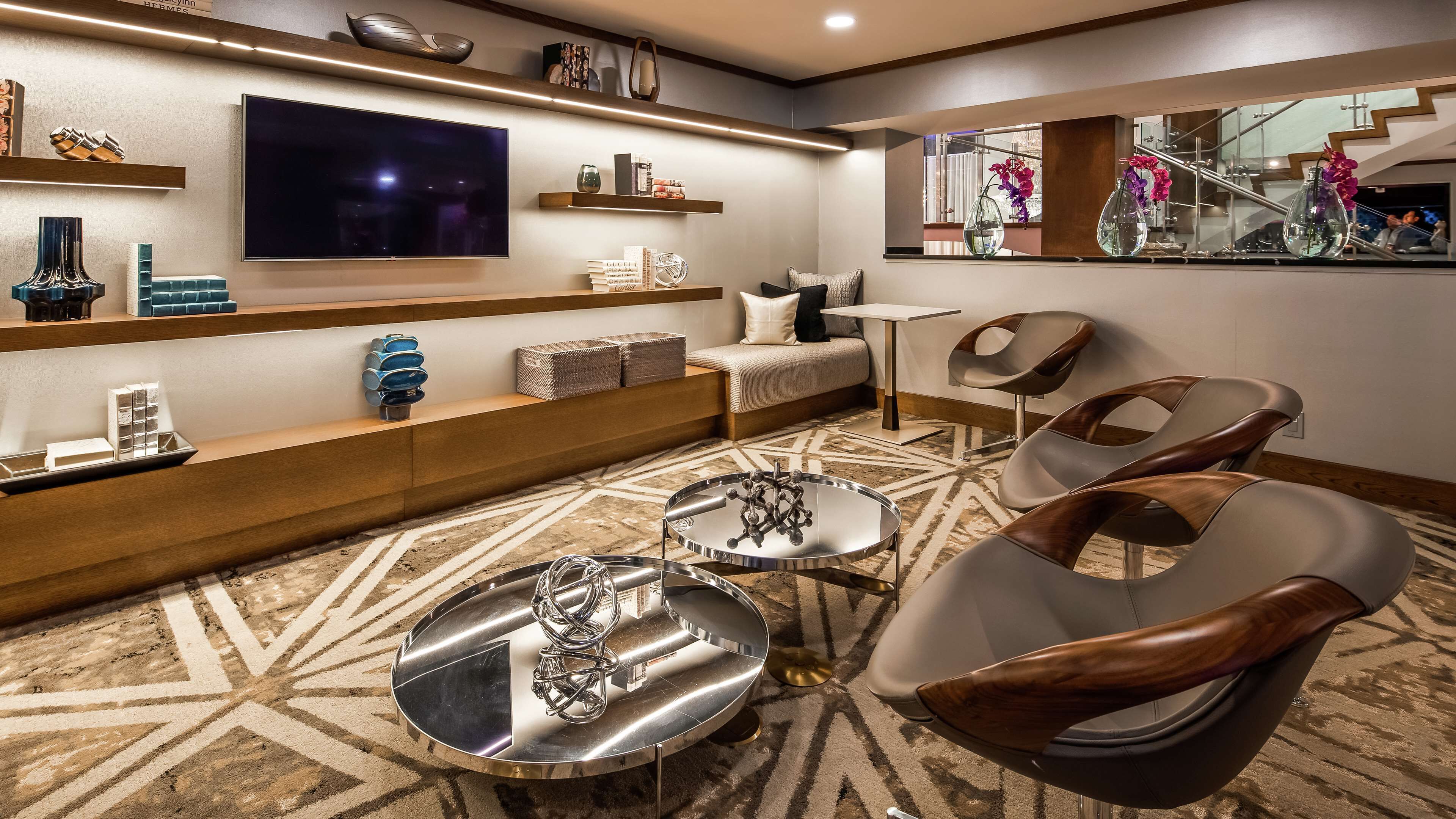 A modern hotel room with a flat-screen TV, a sofa, two chairs, a coffee table, and a patterned carpet.