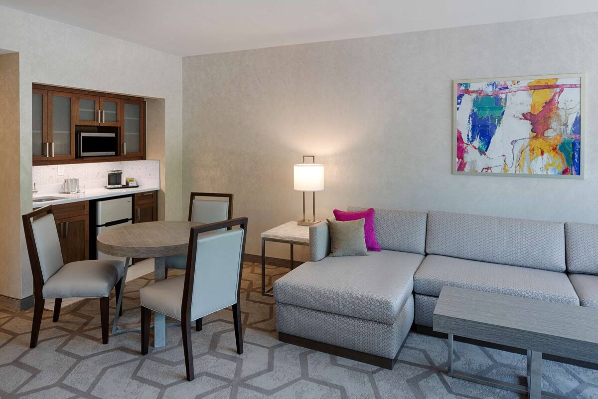 A modern living room with a grey sofa, a dining table with chairs, a kitchen area with wooden cabinets, and a colorful abstract painting on the wall.