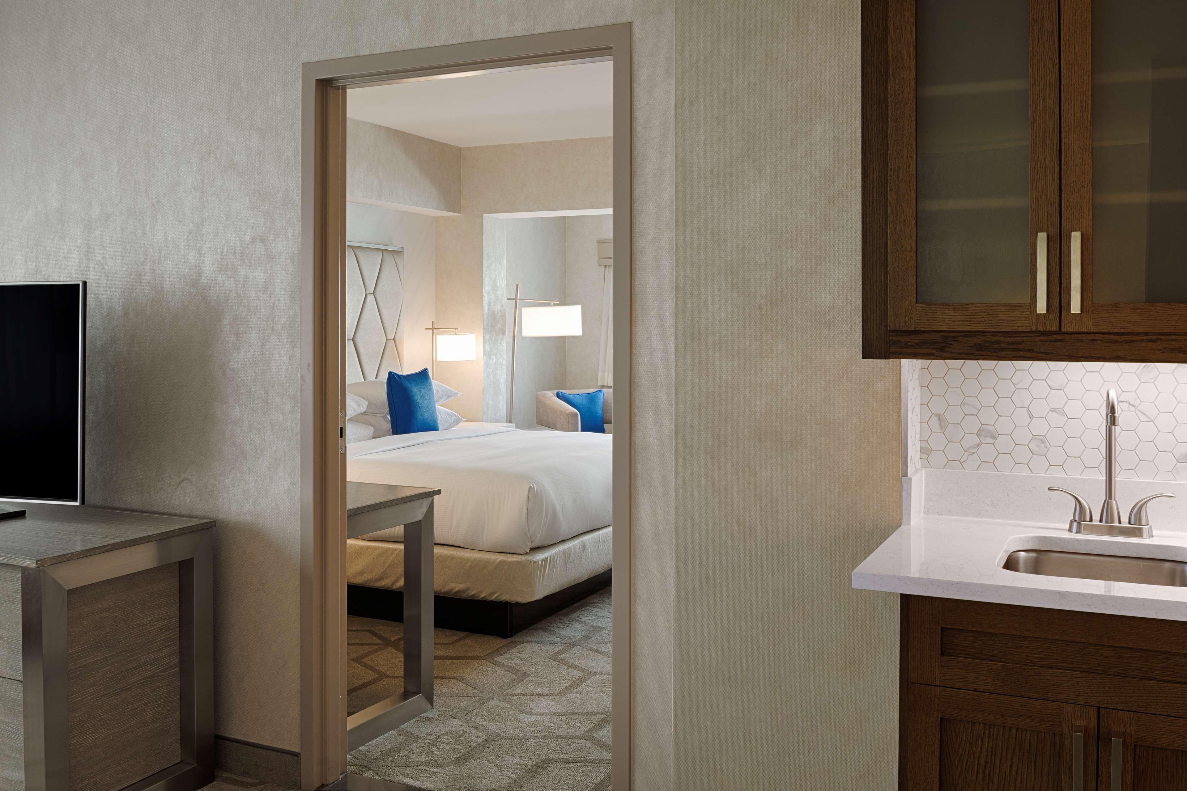 Habitación de hotel con cama, mesita de noche y televisor. Incluye un baño con lavamanos y espejo.