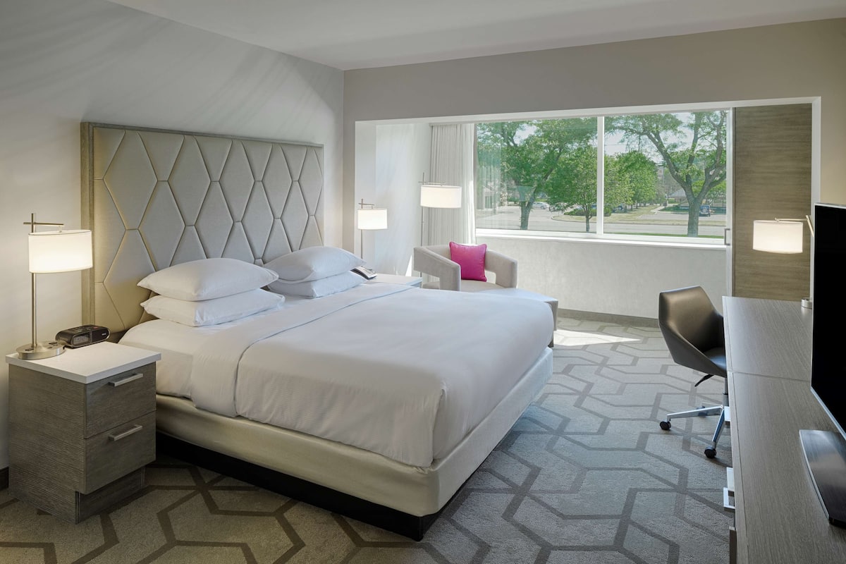 A hotel room with a large bed, bedside tables, a desk, and a chair. There is a window with a view of the outside.