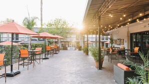 An outdoor patio area with tables and chairs under umbrellas, surrounded by potted plants and a tall palm tree. - Palm Garden Hotel (Thousand Oaks)