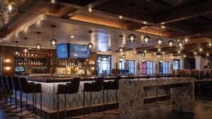 A bar area with a marble counter, multiple bar stools, hanging spherical light fixtures, and a television screen displaying "Selwyns". - Palm Garden Hotel (Thousand Oaks)
