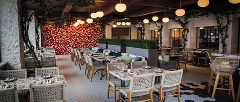 Un restaurante con mesas largas, sillas de mimbre, lámparas colgantes y una pared cubierta de flores rojas.