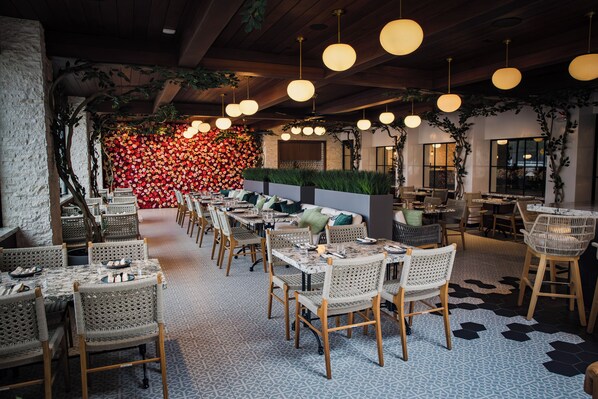 A restaurant with long tables, wicker chairs, hanging pendant lights, and a wall of red flowers. - Palm Garden Hotel (Thousand Oaks)