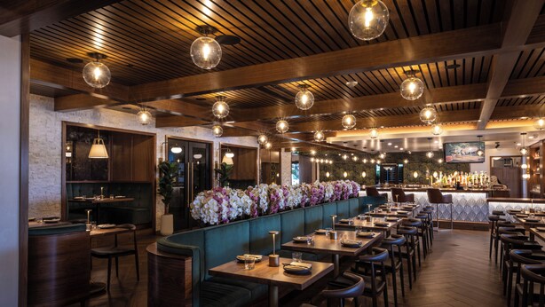 A restaurant with wooden ceiling beams, round pendant lights, and a long counter with a floral arrangement. - Palm Garden Hotel (Thousand Oaks)