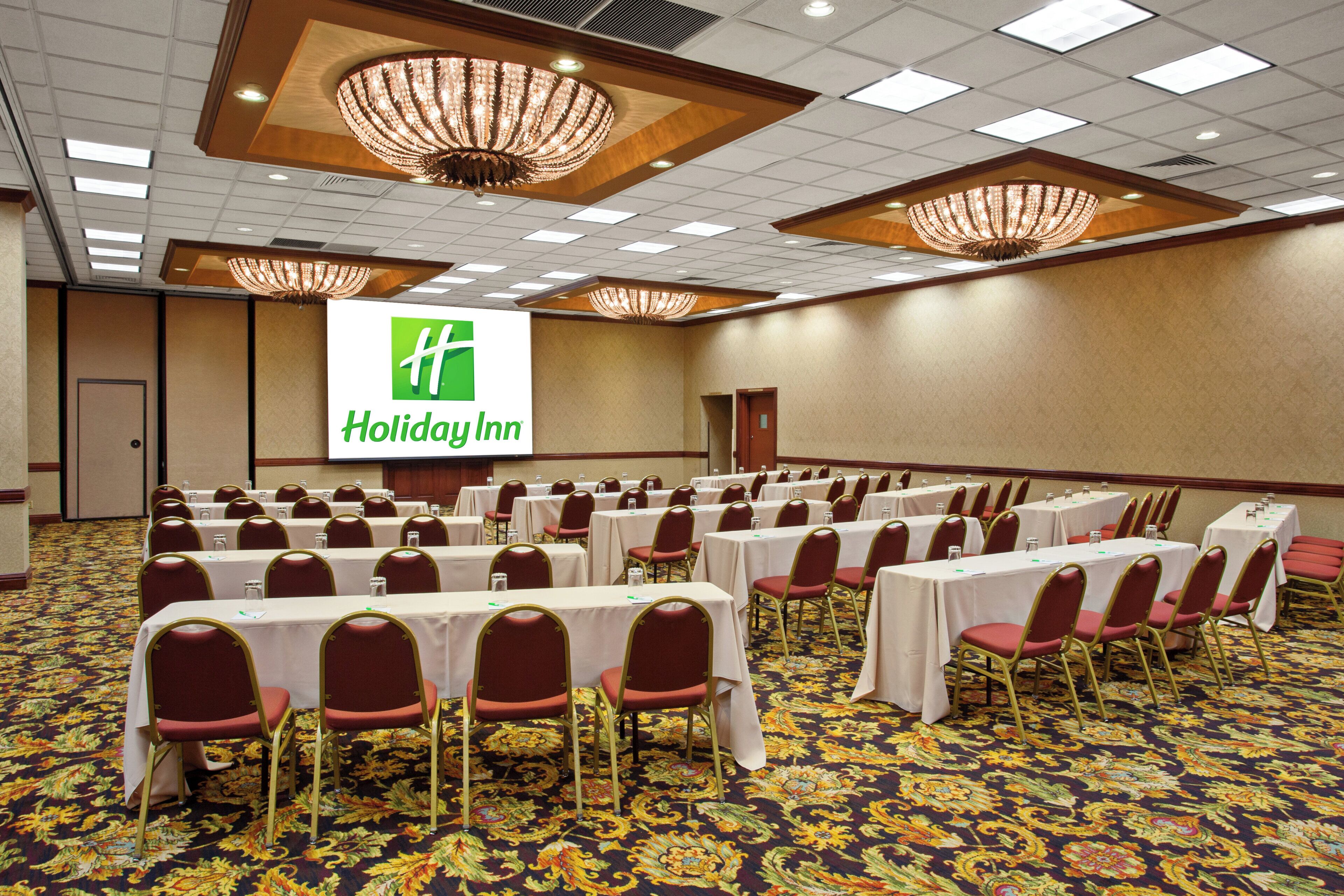 Un salón de conferencias con filas de mesas y sillas, una pantalla grande mostrando el logotipo de Holiday Inn y candelabros en el techo.