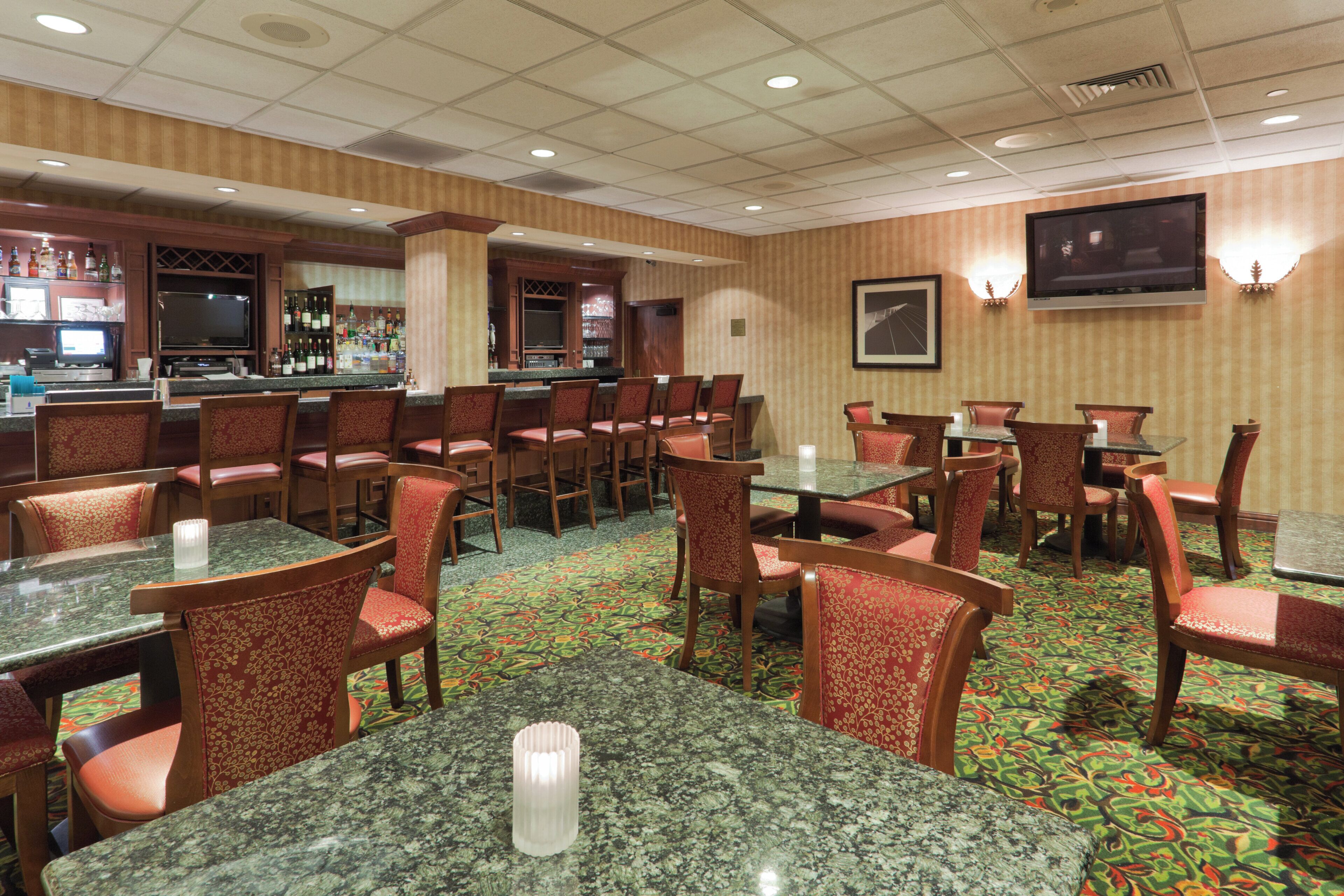 A hotel dining area with multiple tables and chairs, a bar area with bottles and glasses, and a television mounted on the wall.