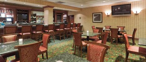 A hotel dining area with multiple tables and chairs, a bar area with bottles and glasses, and a television mounted on the wall.