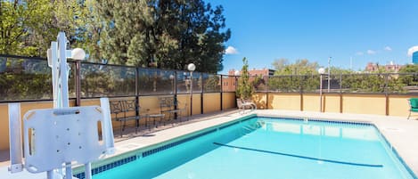 A swimming pool with a safety chair, a table, and chairs, surrounded by a fence and trees.