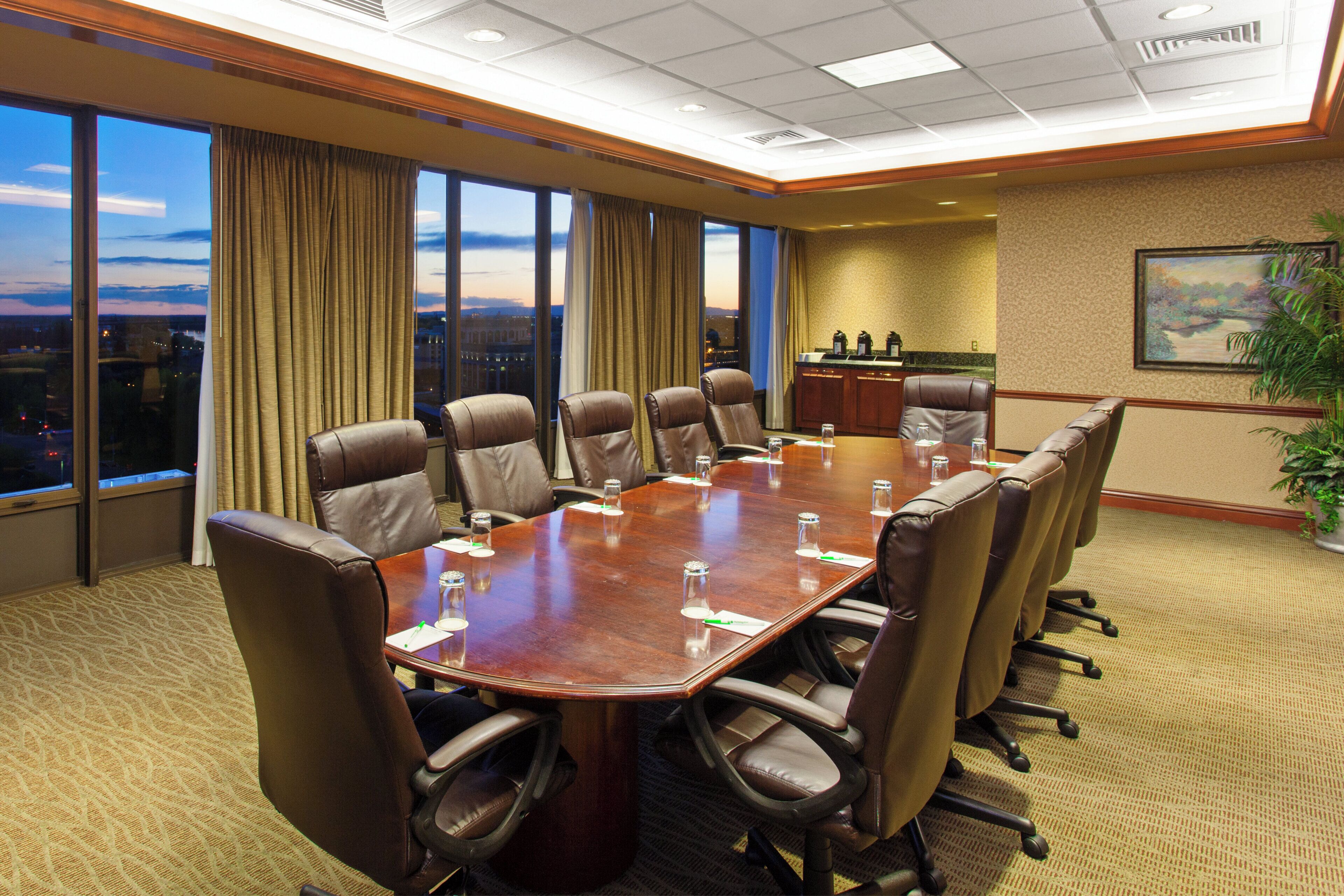 A conference room with a large table, multiple chairs, a view of the cityscape through large windows, and a painting on the wall.