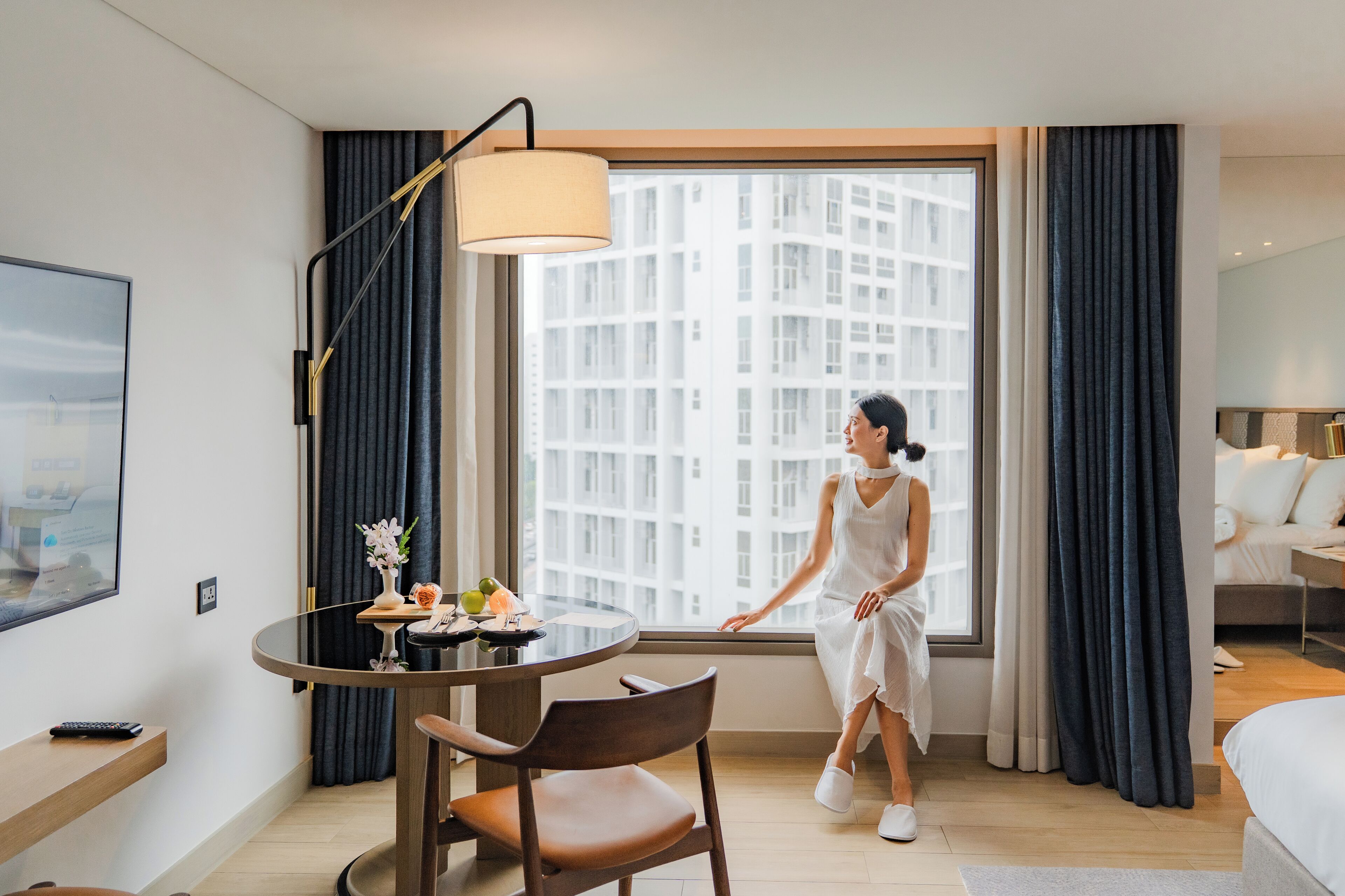 A hotel room with a large window, a round dining table with chairs, a remote control, and a bed.