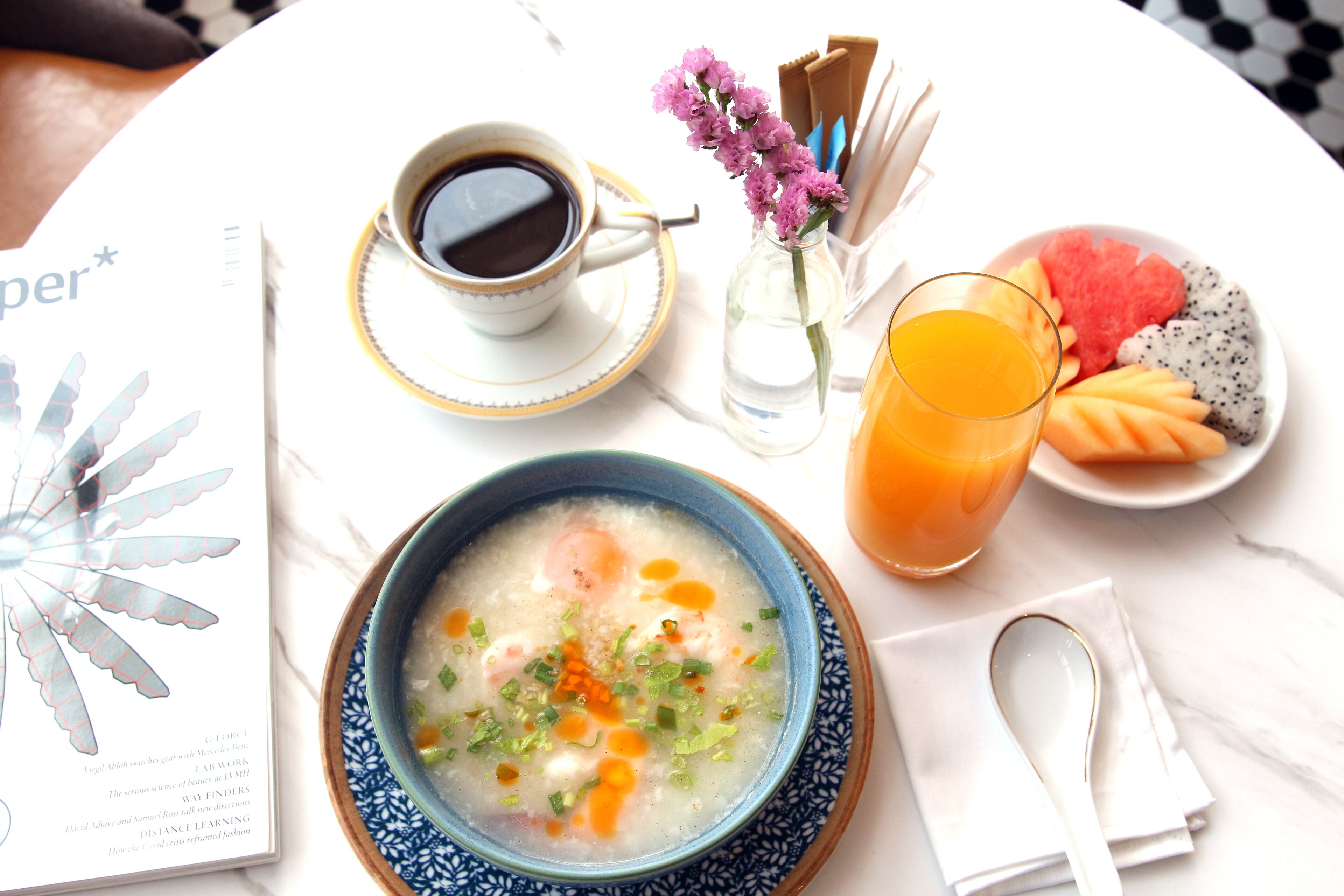 A table with a bowl of soup, a cup of coffee, a glass of orange juice, a plate of fruit, a book, and a vase with flowers.