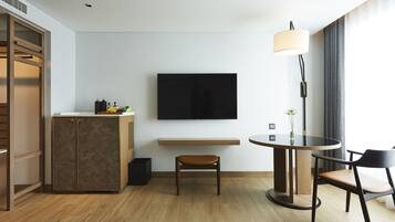 Una habitación de hotel moderna con piso de madera, una mesa de comedor redonda con dos sillas, un televisor de pantalla plana montado en la pared, un mueble bajo con espejo y una lámpara de noche.
