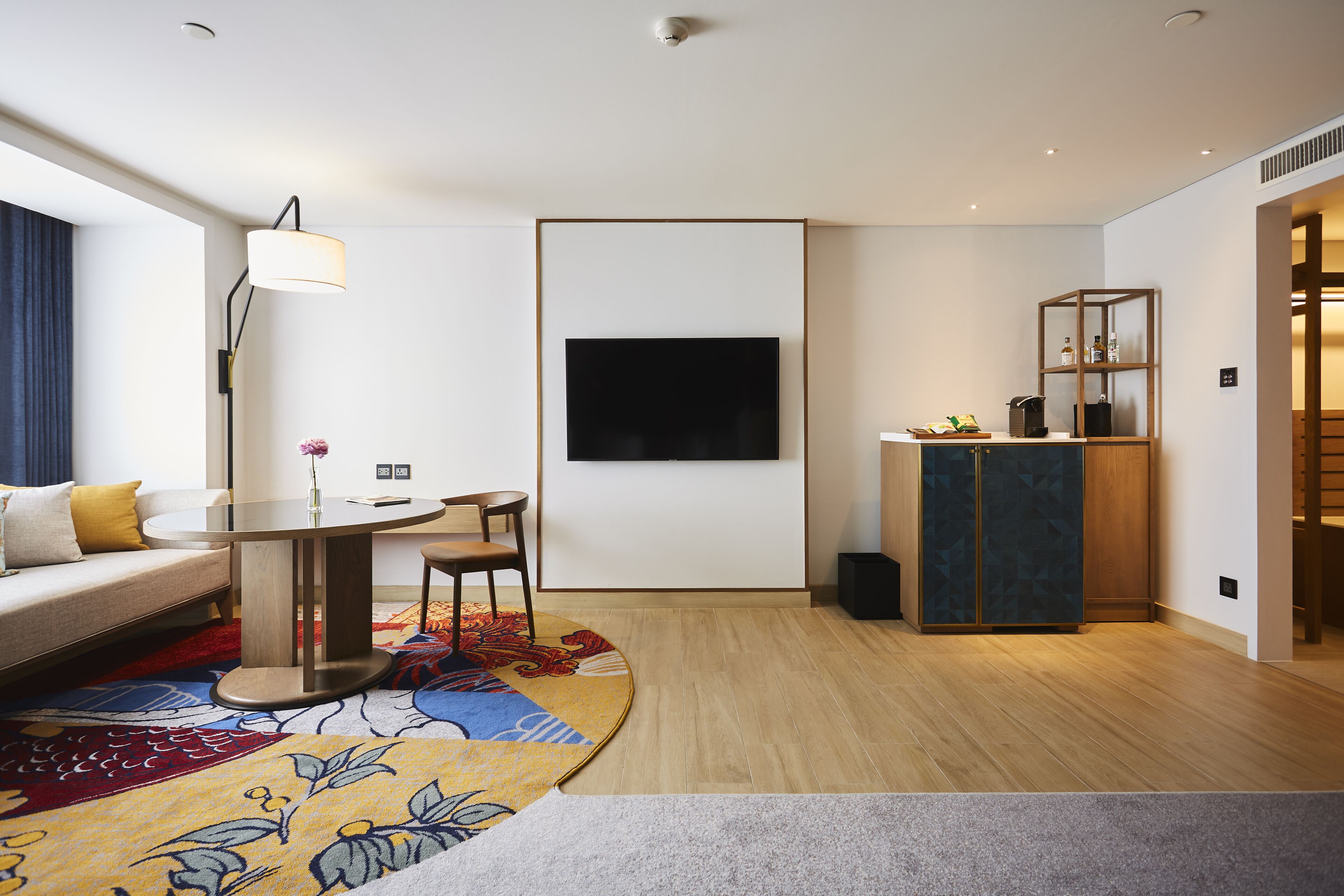 A modern hotel room with a sofa, a dining table, a TV mounted on the wall, a small cabinet, and a patterned rug.