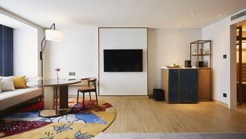 A modern hotel room with a sofa, a dining table, a TV mounted on the wall, a small cabinet, and a patterned rug.