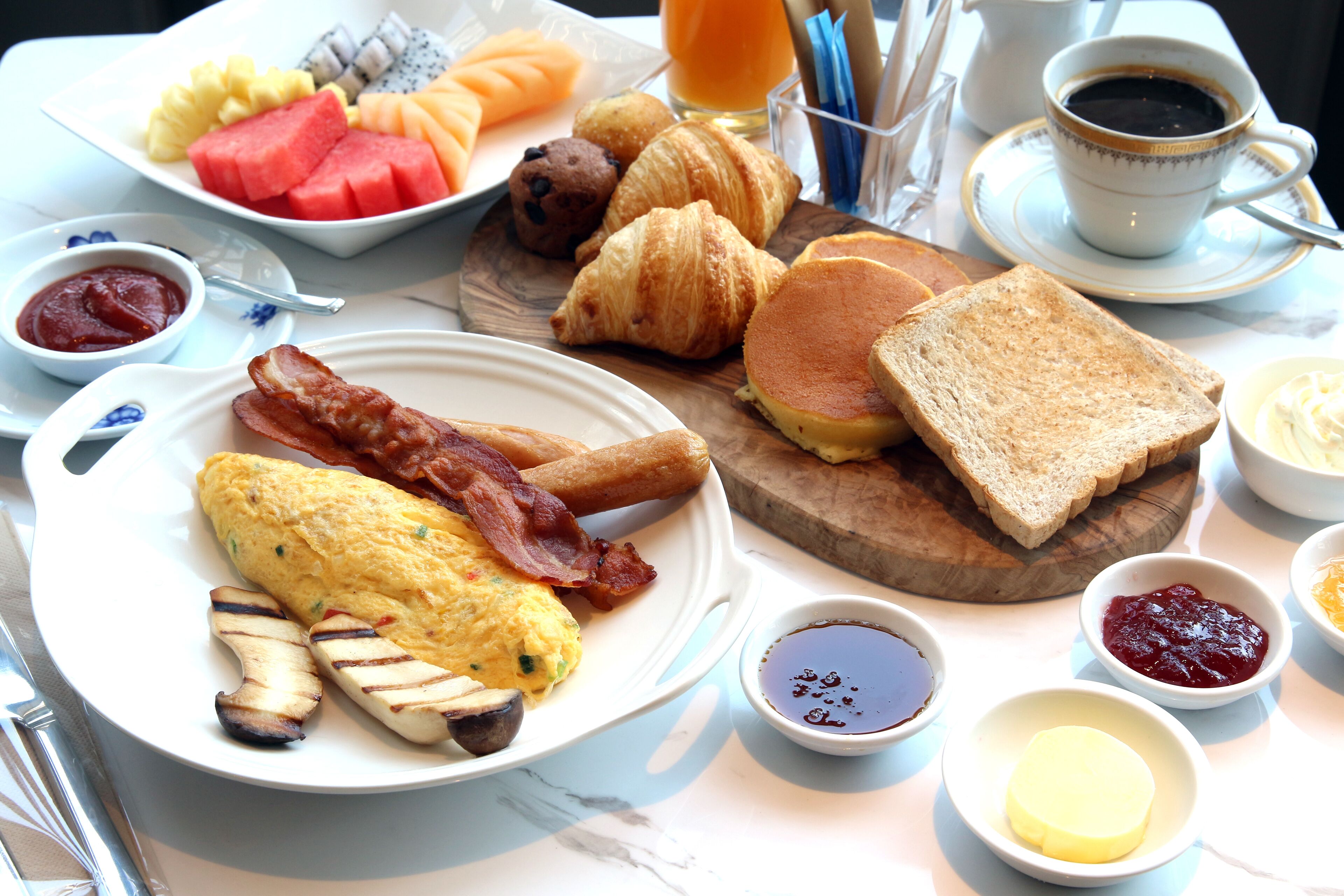 Un desayuno abundante con huevos, tocino, tostadas, croissants y diversas frutas y salsas.