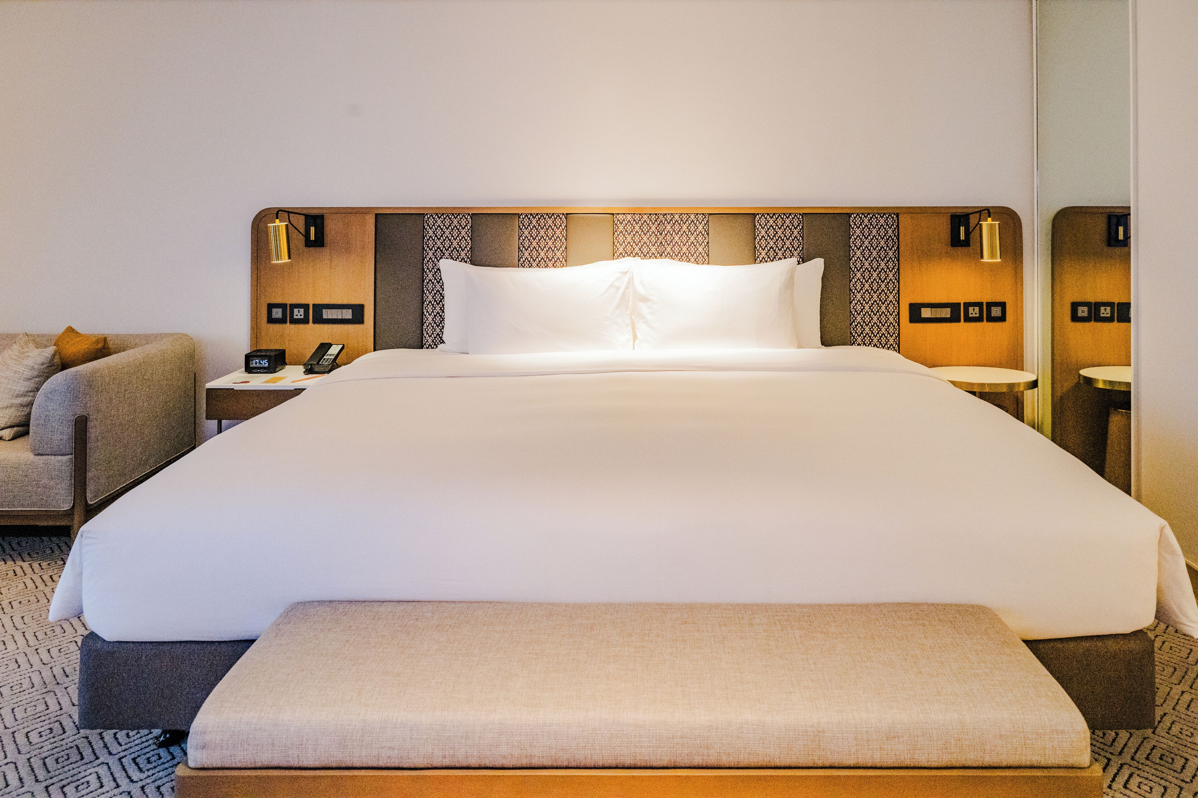 A hotel room with a large bed, a bedside table with a lamp, a chair, and a small table with a phone.
