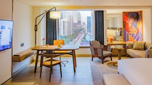 A hotel room with a dining area, a sofa, a TV, and a large window with a city view. - Montien Hotel Surawong Bangkok (Bangkok)