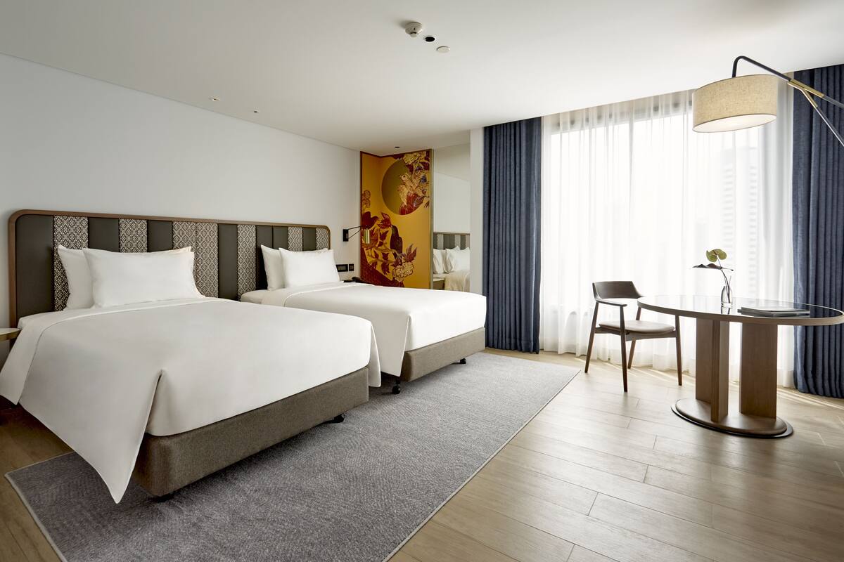 A hotel room with two beds, a desk, a chair, and a large window with curtains.