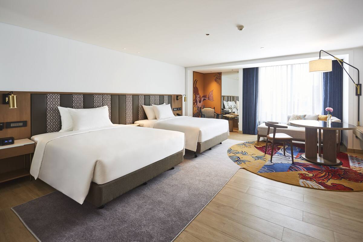 A hotel room with two beds, a desk, a chair, a lamp, and a large window with curtains.