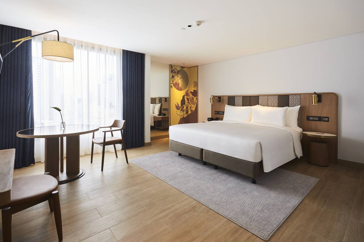 A hotel room with a bed, a round dining table with chairs, a lamp, and a large window with curtains.