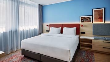 Une chambre d’hôtel moderne avec un grand lit, des tables de chevet en bois, un bureau et une fenêtre agrémentée de rideaux légers.