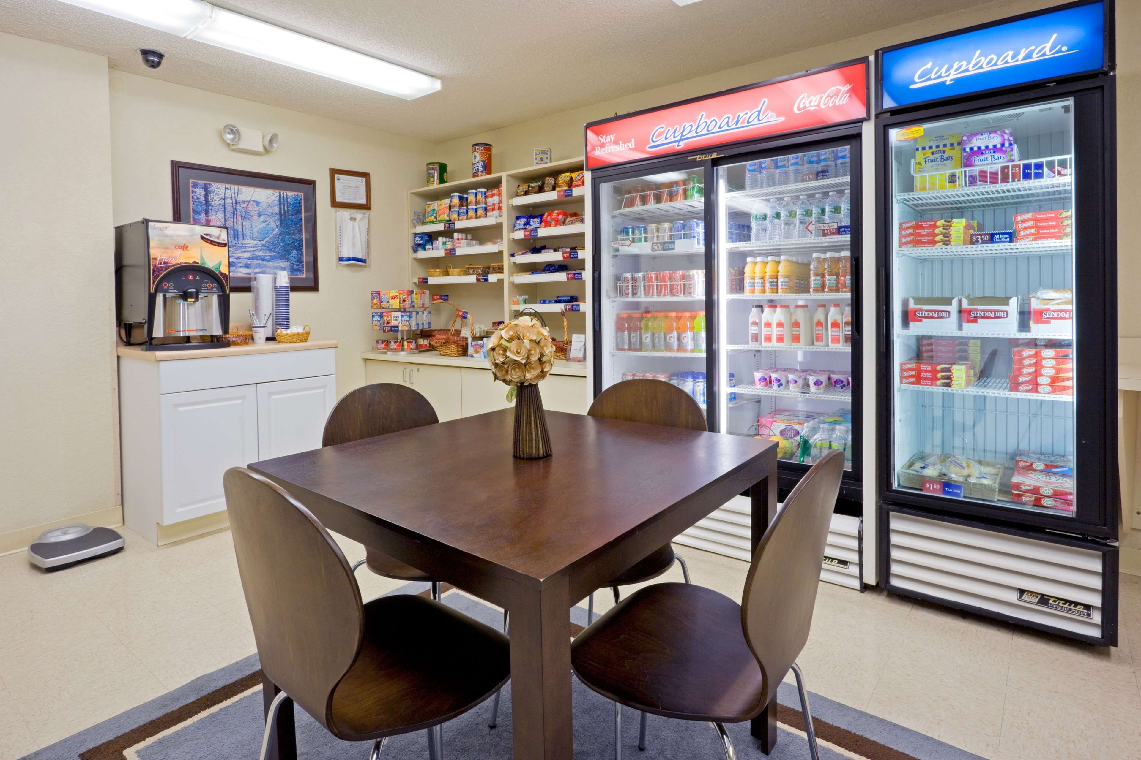 A hotel room with a dining table, chairs, a coffee machine, a scale, a refrigerator, and shelves stocked with snacks and beverages.