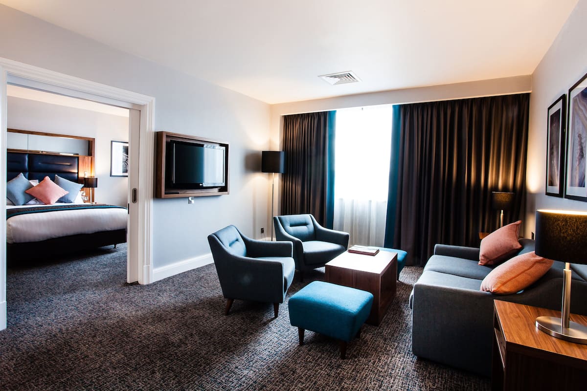 A hotel room with a bed, two armchairs, a sofa, a coffee table, a TV, and a large window with curtains.