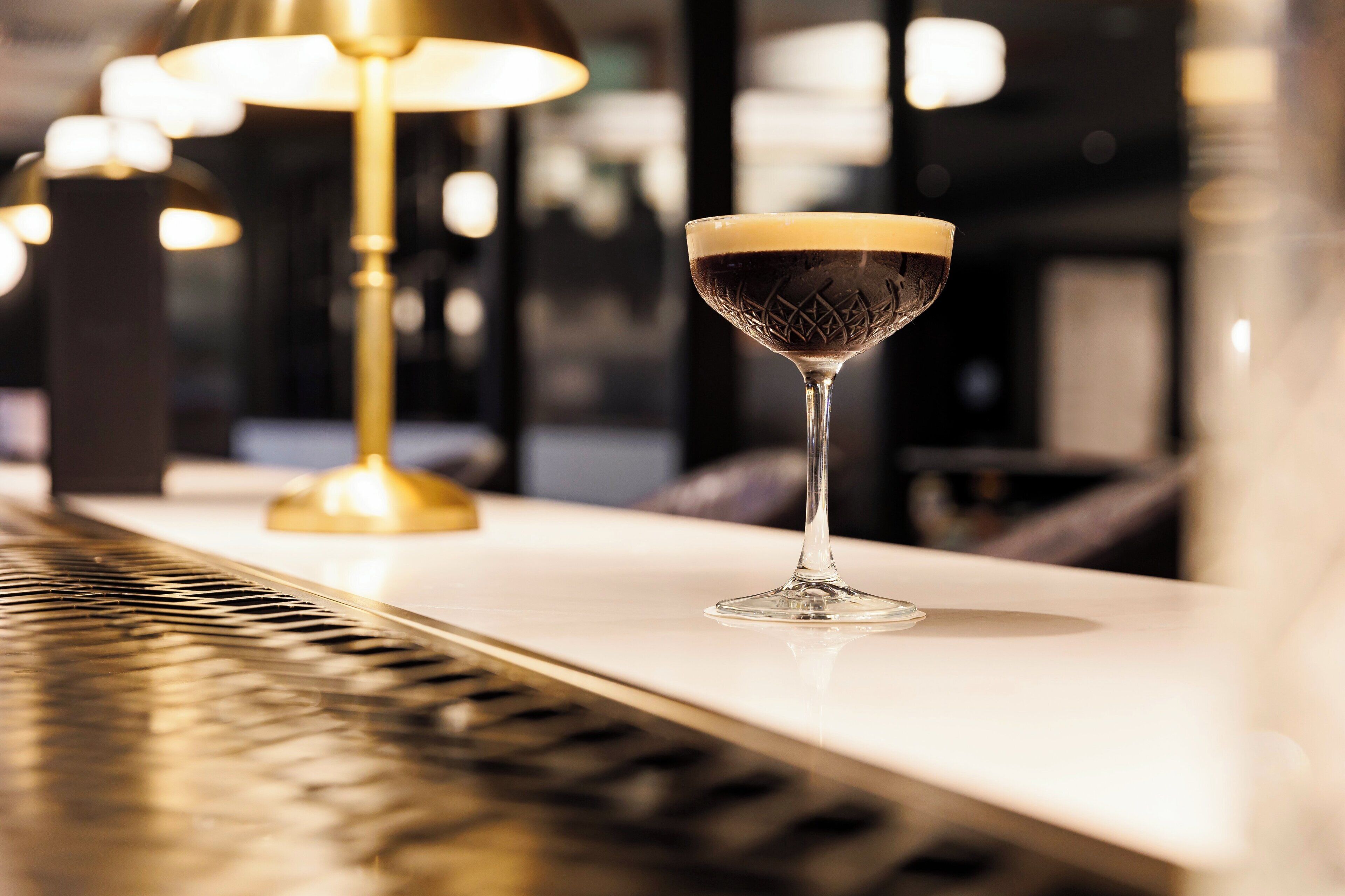 Un verre à martini contenant un liquide sombre, posé sur une surface blanche, une lampe dorée et un arrière-plan flou avec des lumières.