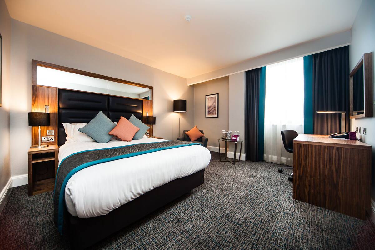 A hotel room with a large bed, a desk, a chair, a lamp, and a view of the city.