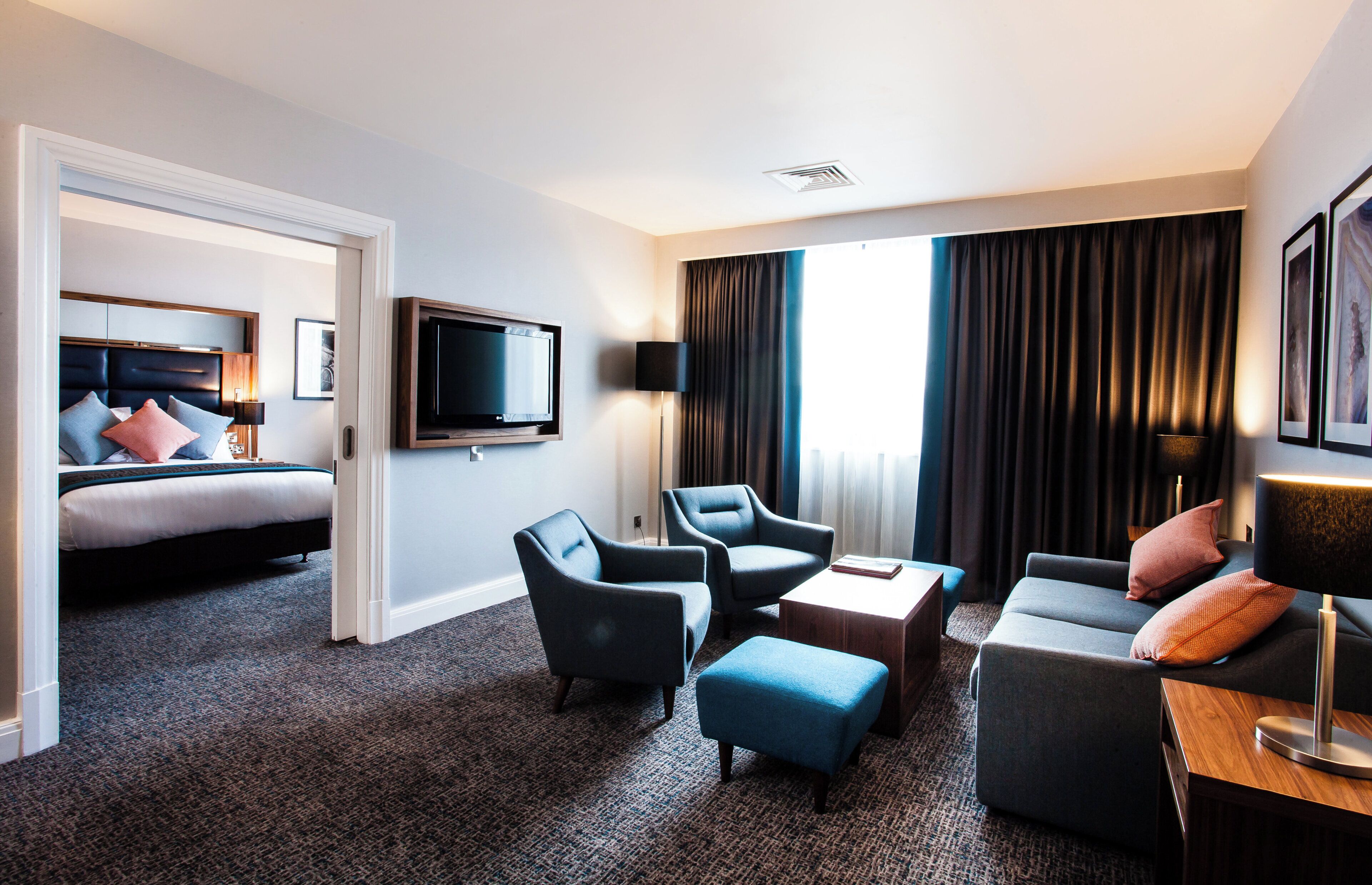 Habitación de hotel con cama, dos sillones, un sofá, televisión y un amplio ventanal con cortinas.