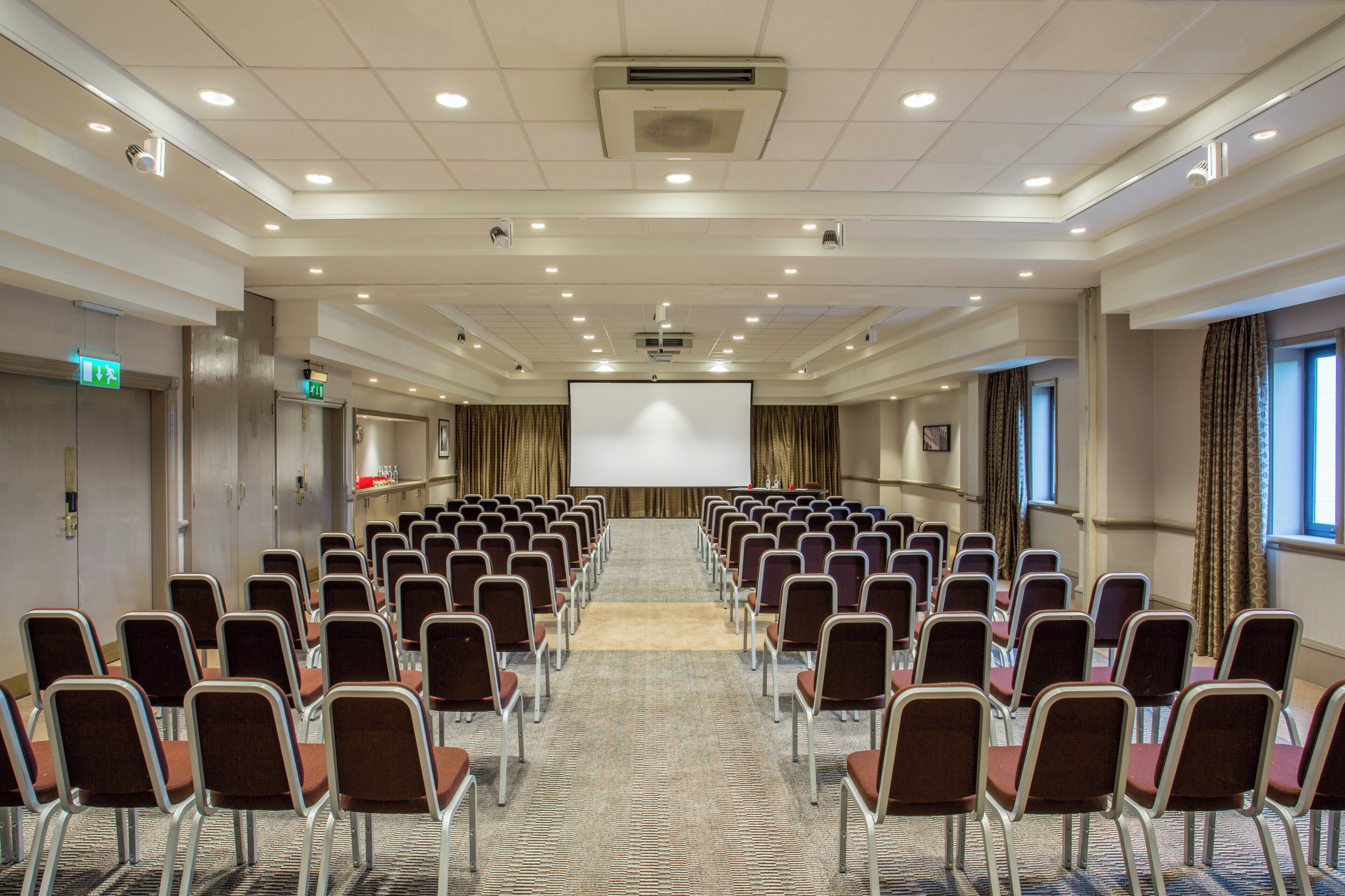 Un salón de conferencias con filas de sillas orientadas hacia una pantalla de proyección, un atril y una señal de salida.