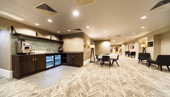 A hotel lobby with a kitchen area, a dining table with chairs, and a patterned carpet.