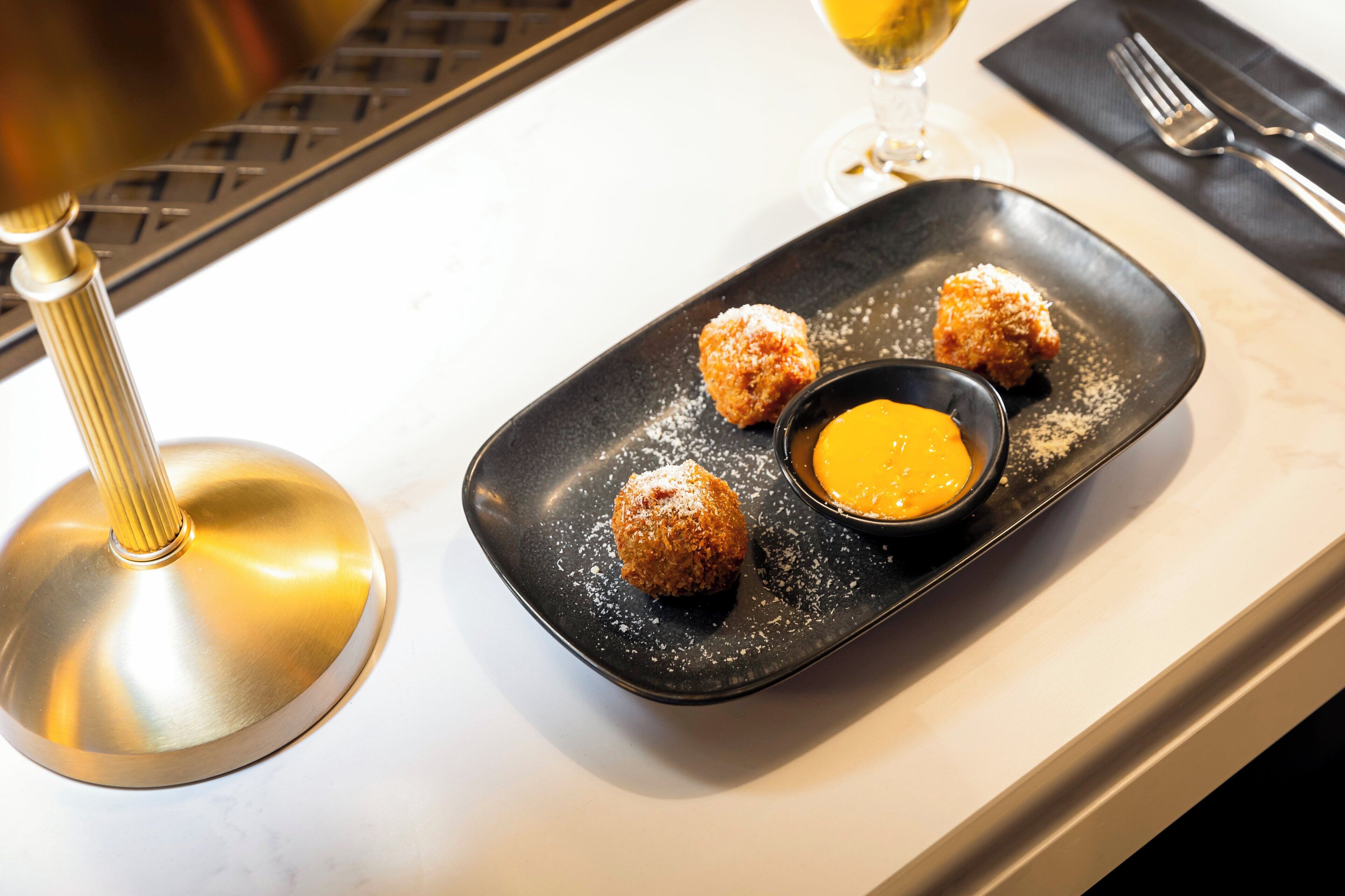 A plate with three fried balls and a small bowl of sauce on a white table.