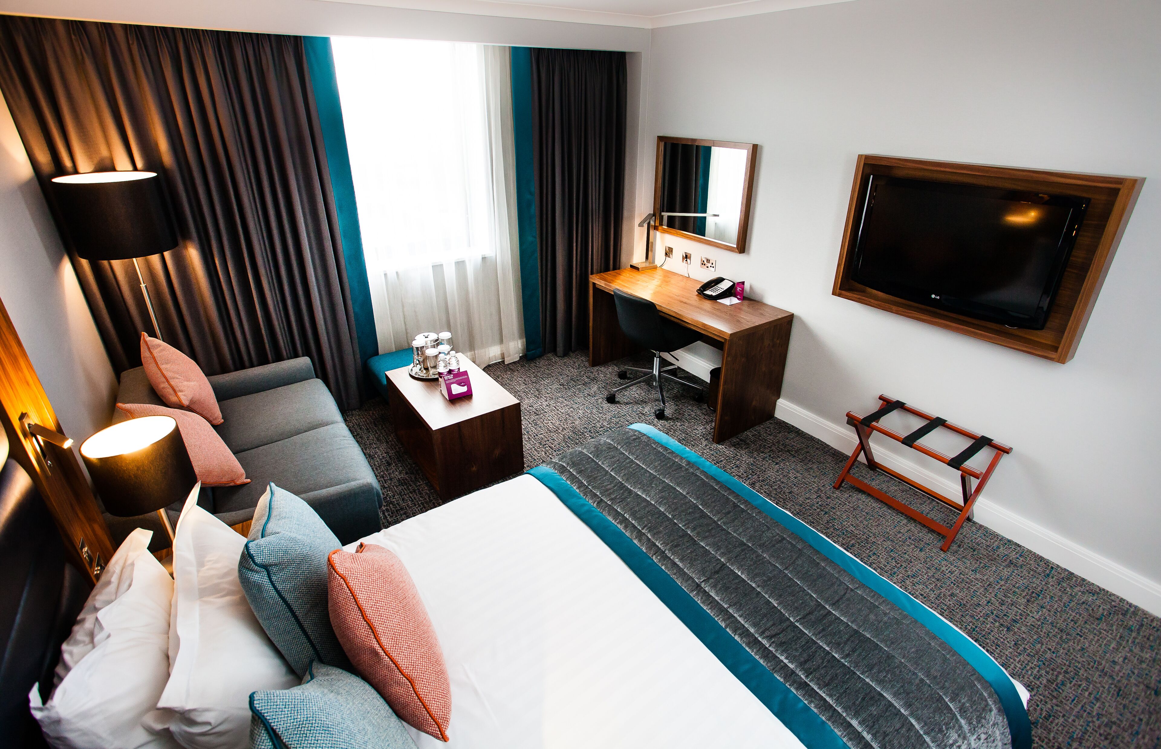 A hotel room with a bed, a desk, a chair, a sofa, a lamp, a TV, and a window with curtains.