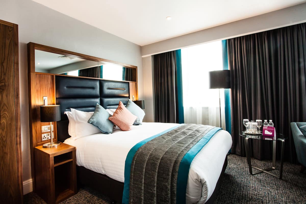 A hotel room with a large bed, a nightstand with a lamp, a small table with a tray, and a chair.