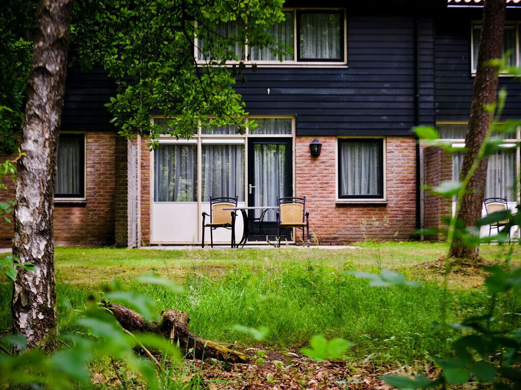 Een gebouw met twee verdiepingen, een bakstenen gevel en grote ramen. Er is een patio met tuinmeubilair.