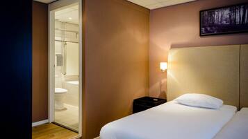 Une chambre d’hôtel comprenant un lit, une table de chevet, une lampe fixée au mur et une porte donnant accès à une salle de bain.