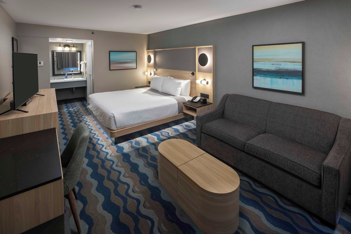 Habitación de hotel con cama, sofá, escritorio, televisión, baño y obras de arte en las paredes.