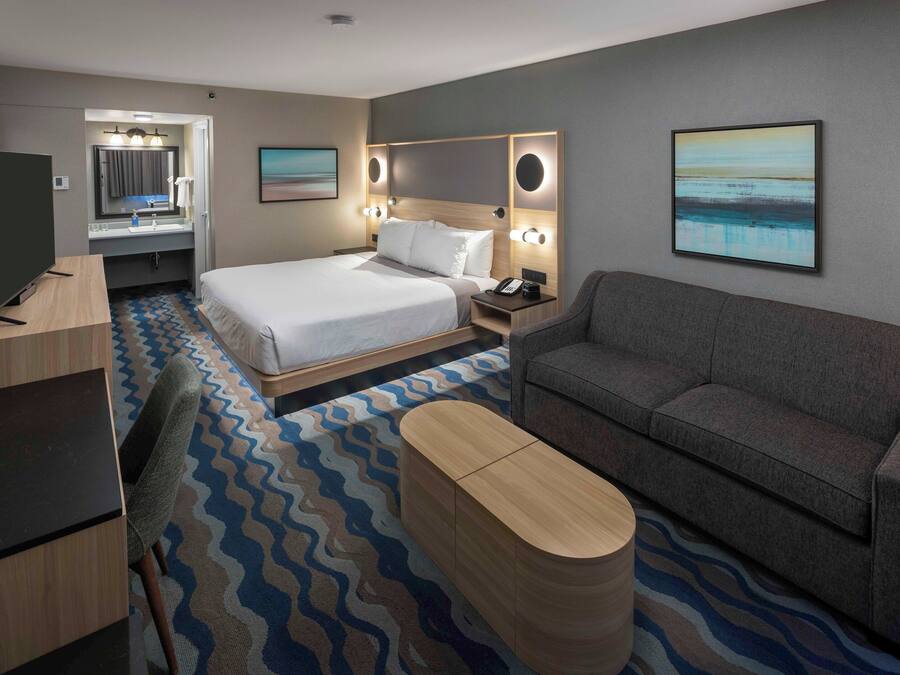 A hotel room with a bed, a sofa, a desk, a television, a bathroom, and artwork on the walls.