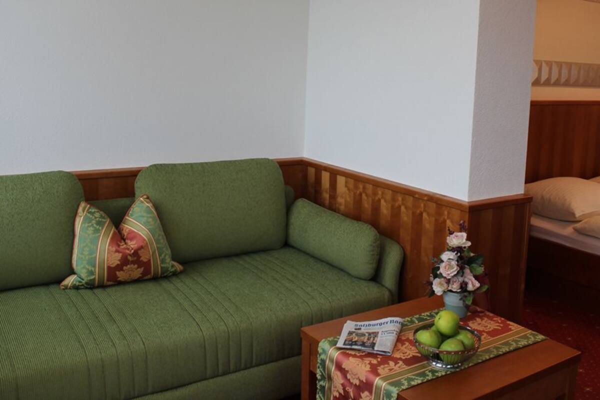 Une pièce avec un canapé vert, un lit avec une tête de lit en bois, une petite table sur laquelle se trouve un bol de pommes et un vase de fleurs, et un magazine posé sur la table.