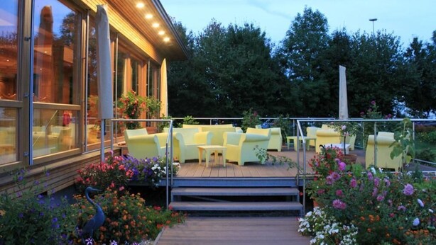 A modern outdoor seating area with yellow chairs and a wooden deck, surrounded by colorful flowers and a peacock statue. - Airporthotel Salzburg (Salzburg)