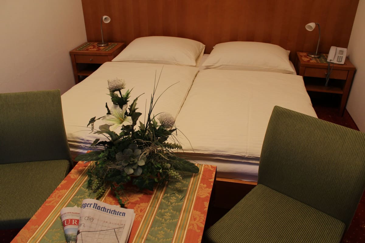 Une chambre d’hôtel avec un lit, deux tables de chevet avec des lampes, une petite table avec une plante et un magazine, et deux chaises vertes.