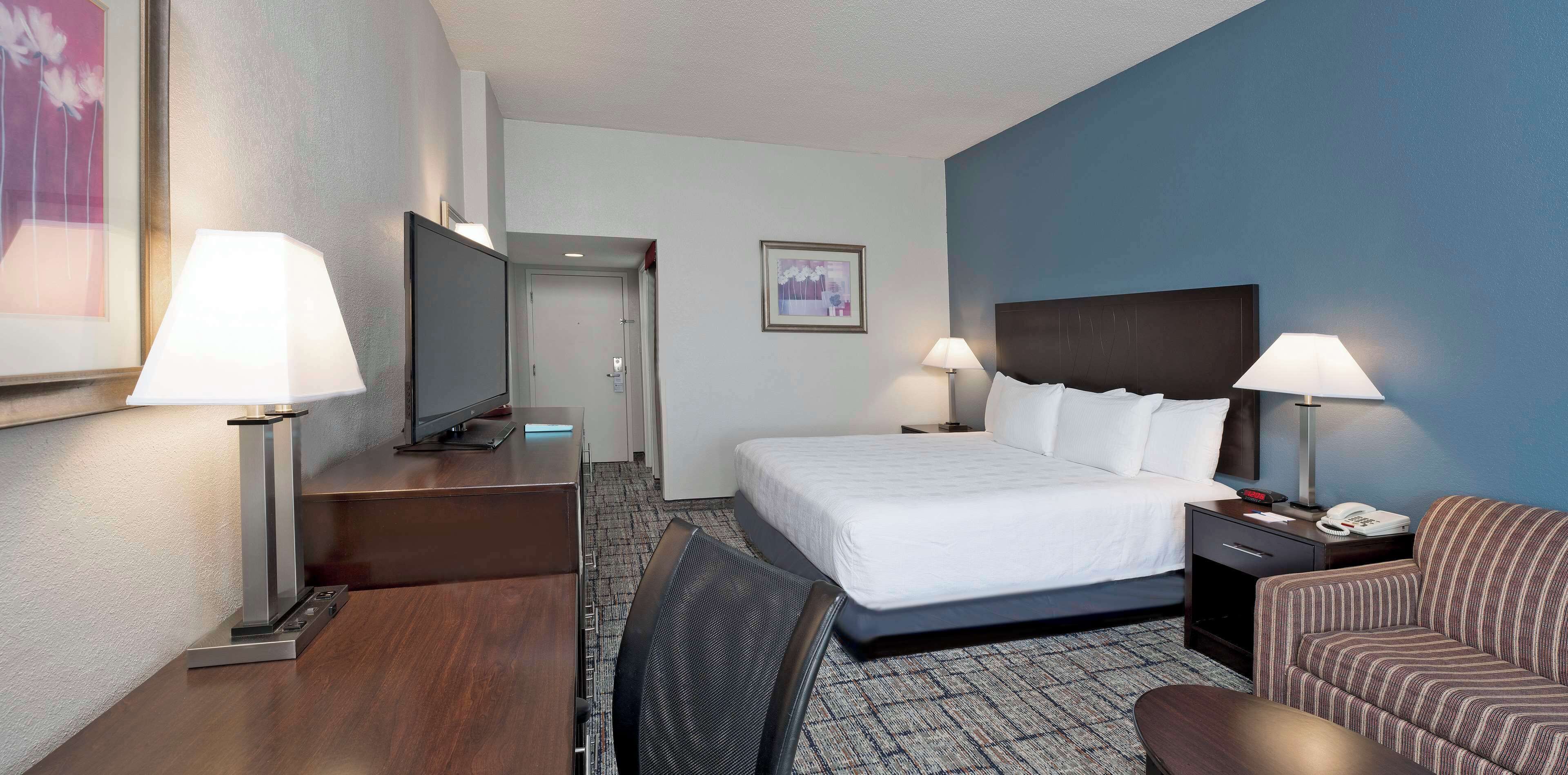 Une chambre d’hôtel comprenant un lit, deux tables de chevet avec des lampes, un bureau avec un ordinateur, une télévision, une chaise et un sol recouvert de moquette.