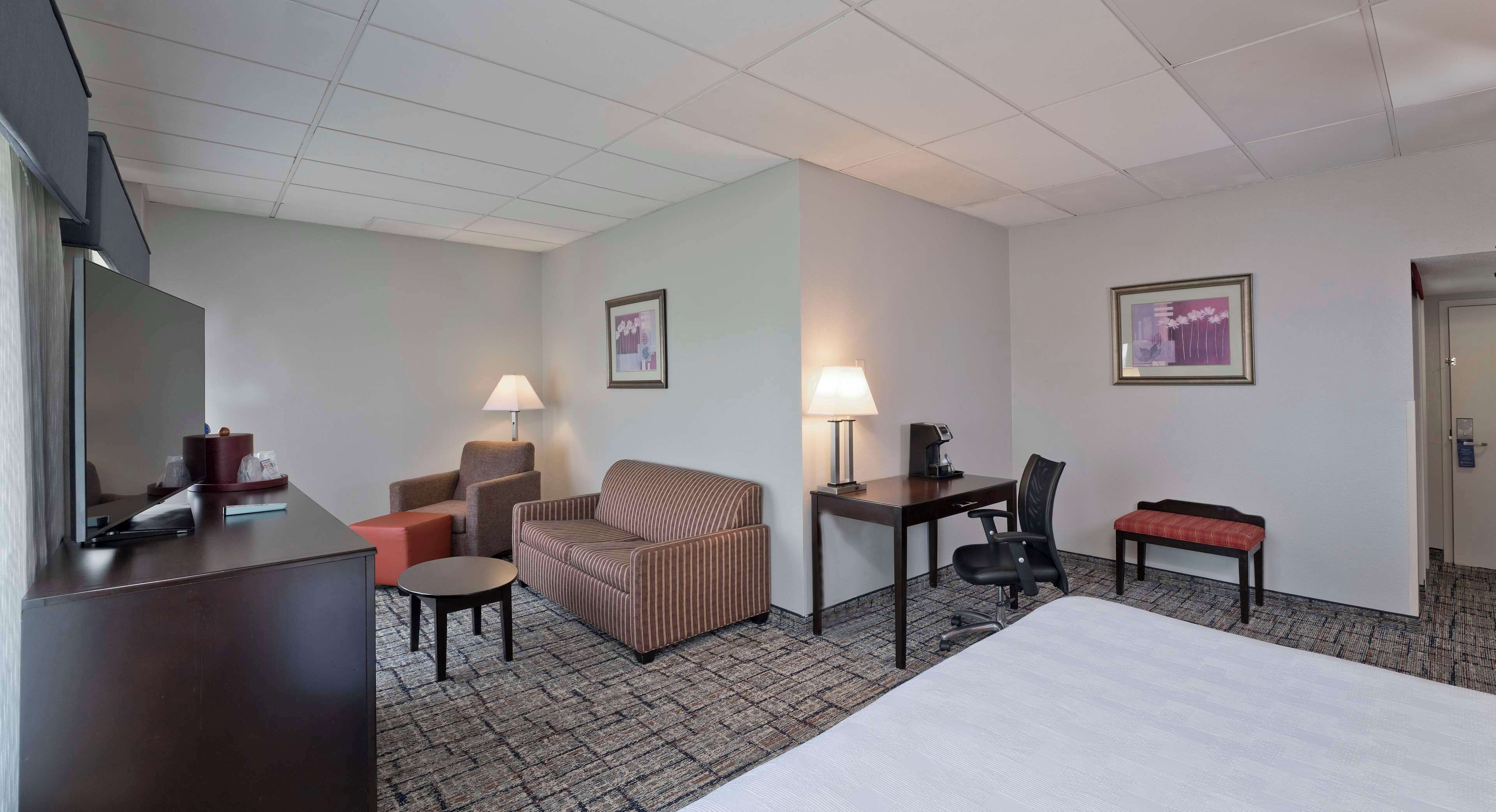 Habitación de hotel con cama, escritorio con silla, sofá, mesita pequeña, lámpara y televisor.
