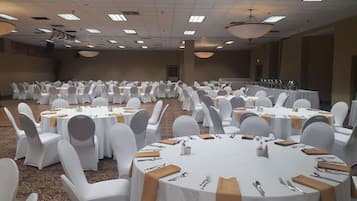 Un salón de banquetes con mesas redondas dispuestas para comer, sillas blancas e iluminación colgante.