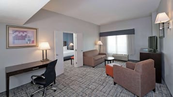 Ett hotellrum med ett skrivbord, två stolar, en soffa, en säng, en tv och ett fönster med gardiner.