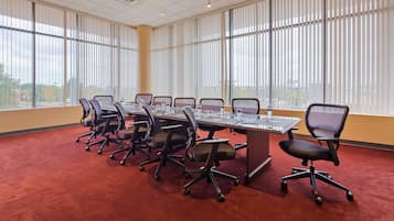 Un salón de conferencias con una larga mesa, varias sillas y grandes ventanales con persianas.