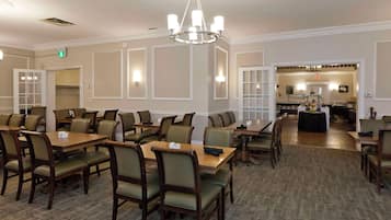 A hotel dining area with multiple tables and chairs, a chandelier, and a kitchen in the background.